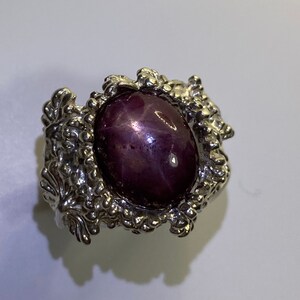 May include: A silver ring featuring a large, oval-shaped, purple star ruby gemstone set in an intricate, floral design.