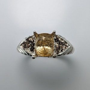 Puede incluir: Un anillo de plata con una gran piedra preciosa cuadrada de color amarillo dorado con líneas internas. El anillo tiene piedras preciosas más pequeñas de color marrón oscuro a cada lado de la piedra principal, engastadas en una banda de plata decorativa.