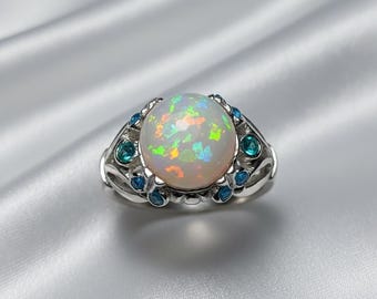 May include: A silver ring featuring a large, round opal gemstone with iridescent colors. The ring is accented with small, round, blue gemstones. The ring is set against a white, silky background.