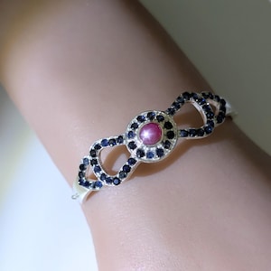 May include: Silver bracelet with a central pink gemstone and small, dark blue stones. The bracelet has an open design with repeating infinity-like shapes, each adorned with dark blue stones. A delicate piece of jewellery.