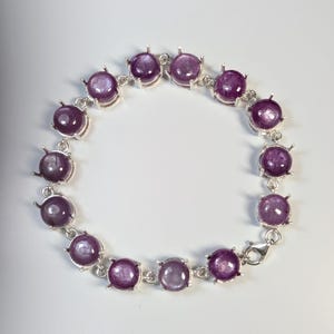 May include: A silver bracelet featuring round, deep purple gemstones. Each gem is set in a silver bezel and connected by silver links. The bracelet is secured with a silver clasp. The gemstones have a polished, reflective surface.