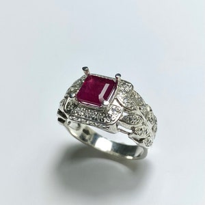 Könnte beinhalten: Ein silberner Ring mit einem roten Edelstein, der von kleineren weißen Edelsteinen umgeben ist. Die Ringschiene ist mit einem Blattdesign verziert.