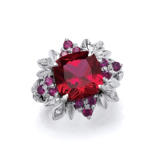 Peut inclure: Une bague en argent ornée d'une grande pierre rouge foncé de taille carrée. De plus petites pierres rondes de couleur rubis et des éléments argentés en forme de feuilles entourent la pierre centrale. De minuscules pierres transparentes sont parsemées dans le design.