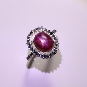 Könnte beinhalten: Ein silberner Ring mit einem großen, ovalen, roten Sternrubin, der von einem Heiligenschein aus kleineren blauen Saphiren umgeben ist.