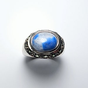 Puede incluir: Un anillo de plata con una piedra ovalada azul y blanca engastada en un engaste de plata. El anillo está decorado con un borde de cuentas.