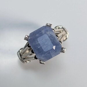Puede incluir: Un anillo de plata con una gran gema rectangular azul claro. El anillo tiene un engaste detallado y ornamentado con pequeños detalles negros. La gema está facetada, captando la luz y mostrando un brillo sutil. El anillo se presenta sobre un fondo blanco.