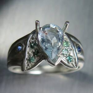 May include: A silver ring with a pear-shaped blue gemstone surrounded by smaller green gemstones. The ring has a unique design with three prongs holding the center stone.