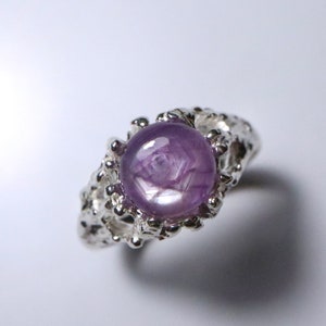 Puede incluir: Un anillo de plata con una piedra preciosa de zafiro estrellado púrpura. El anillo tiene un diseño único y texturizado.