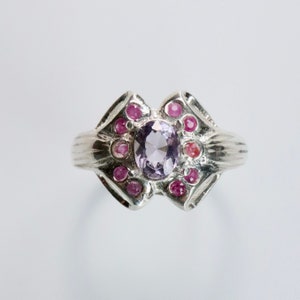 Puede incluir: Un anillo de plata con una piedra ovalada de color púrpura rodeada de piedras pequeñas de color rosa.