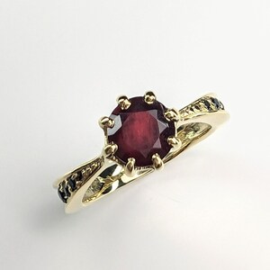 May include: A gold ring featuring a red gemstone set in a crown-like design. The band is adorned with black stones.