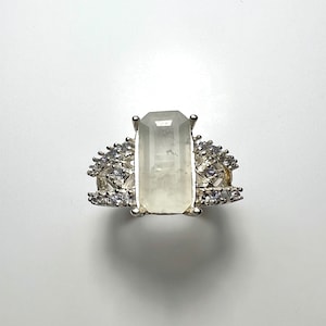 May include: A silver ring featuring a large, clear, rectangular gemstone surrounded by smaller, clear gemstones.