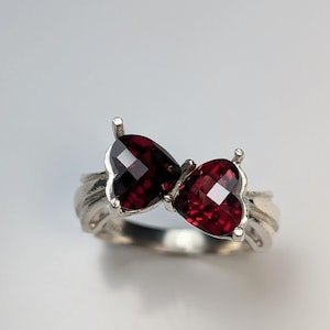 May include: A silver ring featuring two heart-shaped, faceted, deep red gemstones. The ring's band has a textured design, and the gemstones are set close together, creating a bow-like appearance. The ring is displayed against a white background.