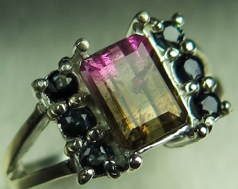 May include: A silver ring with a rectangular, multi-colored gemstone surrounded by smaller black gemstones.