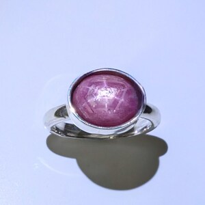 May include: A silver ring with a large, oval star ruby gemstone. The ruby is a deep pinkish-red colour, displaying a star effect. The ring has a simple, polished band and a bezel setting, a classic design.