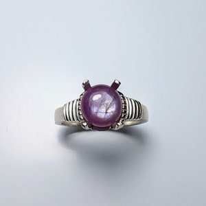 Puede incluir: Un anillo de plata con una gran gema redonda de color rojo púrpura. La gema está engastada en un engaste de cuatro puntas, y la banda tiene un diseño acanalado. El anillo se muestra sobre una superficie blanca.