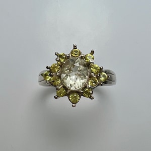 Puede incluir: Un anillo de plata con una gran piedra preciosa ovalada transparente rodeada de pequeñas piedras preciosas amarillas.