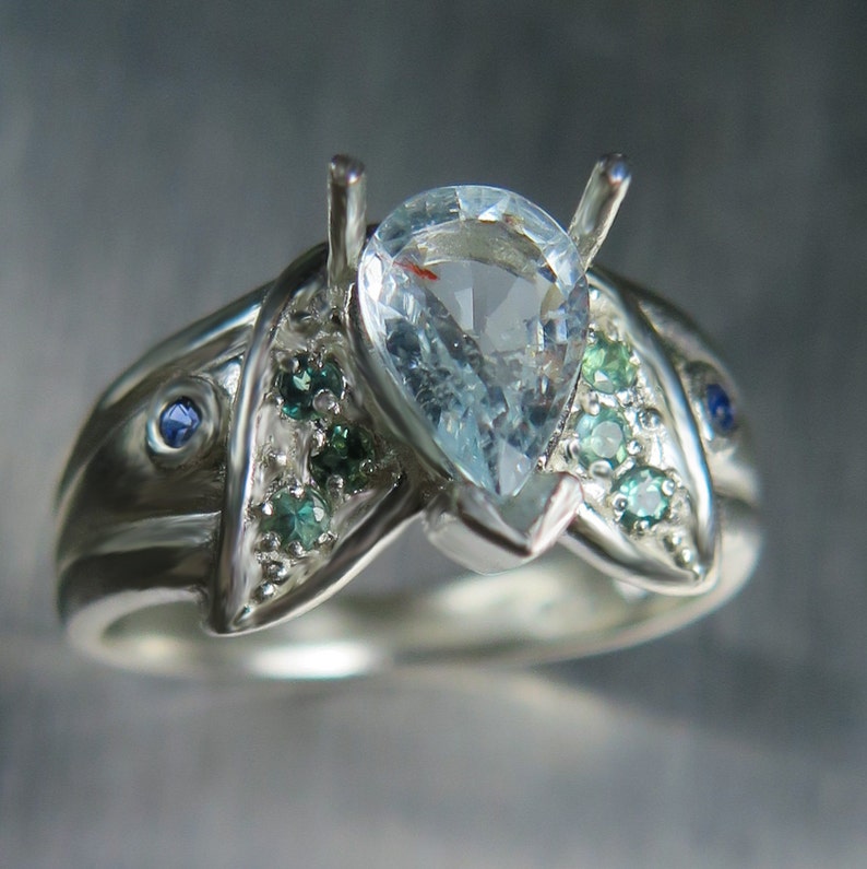 May include: A silver ring with a pear-shaped aquamarine gemstone surrounded by smaller green and blue gemstones.