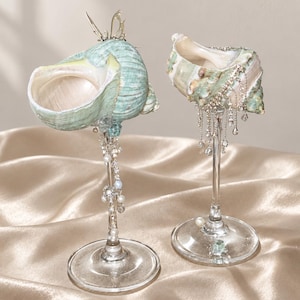 May include: Two decorative goblets featuring sea shells as bowls, colored in shades of mint green and white. The stems are clear glass, adorned with dangling silver chains, small white pearls, and clear crystals. One shell features a small metal butterfly.