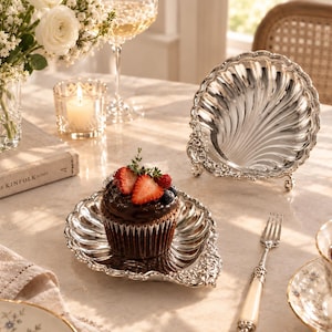 May include: A silver-plated shell-shaped dish with ornate floral detailing along the edges. The dish features a scalloped design and is shown both as a flat plate holding a chocolate cupcake with berries and as an upright display piece.