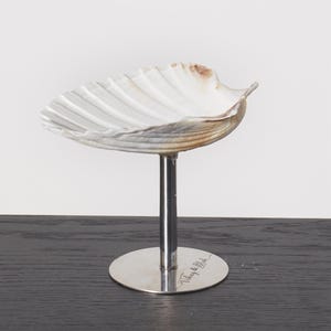 May include: A decorative seashell dish on a silver metal stand. The shell is white with brown and tan markings. The stand has a round base with engraved text.