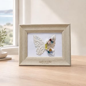 May include: A light wood-toned rectangular frame containing a butterfly art piece. The left wing is textured white, while the right wing is composed of various seashells, pearls, and colourful glass beads. The text Sylvie & Ellis 09.15.2026 is printed.