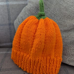 May include: A knitted orange pumpkin hat with a green stem. The hat is made with a soft yarn and has a ribbed texture.