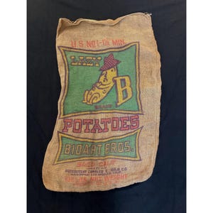 May include: Vintage burlap potato sack with a green and red graphic. The graphic features a cartoon potato wearing a hat, the word "LAZY", and the brand name "BIDART BROS." The sack reads "U.S. NO 1-1/2 MIN" and "100 LBS NET WEIGHT".