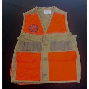 May include: A tan and orange hunting vest with multiple pockets and a quilted orange shoulder patch. The vest has a circular patch that reads "Graduate". The vest has a button-up front and several cartridge holders.