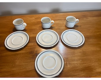 May include: Set of five white ceramic coffee cups and saucers with a thin black trim. The cups and saucers are arranged on a wooden surface.