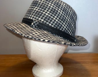 May include: A fedora-style hat with a black and white houndstooth pattern. The hat features a black band around the crown and a structured brim. The hat is displayed on a white mannequin head.