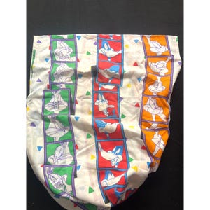 May include: A white fabric sheet featuring vertical film strip patterns containing cartoon character illustrations. The strips are bordered in green, red, and orange, showcasing Bugs Bunny and Daffy Duck. Small colorful triangles are scattered throughout.