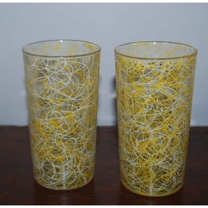 May include: Two clear drinking glasses with a yellow and white abstract design. The glasses are tall and cylindrical, with a pattern of interwoven lines. The glasses are likely vintage and add a retro touch to any kitchen.