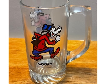 May include: Clear glass mug featuring a colorful image of Goofy in a running pose. Goofy is wearing a red shirt, blue pants, and a black hat. The mug has a handle and a decorative base. The text "GOOFY" and "Walt Disney Productions" are printed on the mug.