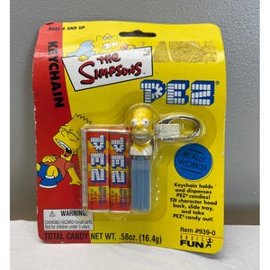 May include: A Simpsons PEZ keychain in original yellow packaging featuring Bart and Maggie graphics. The set includes a Homer Simpson character head dispenser, a metal keyring, and two packs of assorted fruit candy with a total weight of .58 oz.