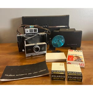 May include: A vintage Polaroid Automatic 250 Land Camera with accessories, including a flash, film, and a case. The camera is black and silver, with the brand name visible. Additional boxes of Polaroid film and flashbulbs are also present.