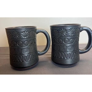 May include: Two black ceramic mugs with decorative floral, circle, and spiral patterns. Each mug has a large handle and a series of embossed designs around the body. The mugs are likely handmade and have a rustic, artisanal feel.