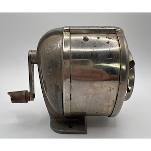 May include: Vintage silver-toned pencil sharpener with a crank handle and a brown knob. The sharpener has a cylindrical design with a base and is likely made of metal. The exterior shows signs of wear, suggesting it's an older model.