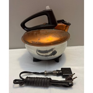 May include: A vintage electric kitchen appliance with a polished silver-tone metal bowl base and a weathered copper-colored lid. It features a black plastic handle, a small spout, and three black feet. A detachable black power cord is included.