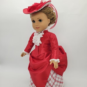 May include: A doll wearing a red satin dress with a white lace collar and cuffs. The dress has a plaid skirt and a matching red hat with a white feather and lace trim. The doll has brown hair and blue eyes.