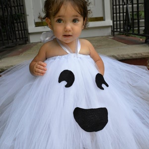 May include: A young child dressed as a ghost for Halloween. The white tulle costume has a black felt ghost face with eyes and a mouth.