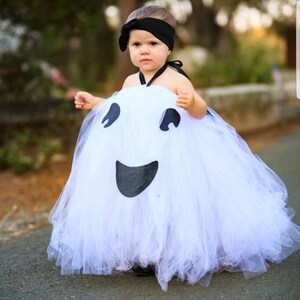 May include: A young child dressed as a ghost for Halloween. The costume is a white tulle tutu with a black ghost face on the front. The child is wearing a black headband.