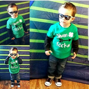 May include: A young child wearing a green t-shirt with the text "Sham Rockin'" and a shamrock graphic. The child is also wearing blue jeans, black sunglasses, and black and white trainers.