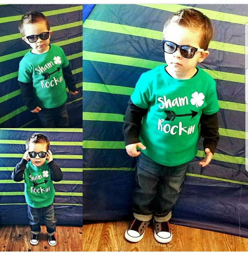 May include: A young child wearing a green t-shirt with the text "Sham Rockin'" and a shamrock graphic. The child is also wearing blue jeans, black sunglasses, and black and white trainers.