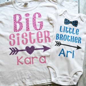 Pu&ograve; includere: Due magliette bianche con un design rosa e blu. La maglietta pi&ugrave; grande dice "Sorella Maggiore Kara" con un cuore glitterato viola e una freccia. La maglietta pi&ugrave; piccola dice "Fratello Minore Ari" con un papillon blu e una freccia.