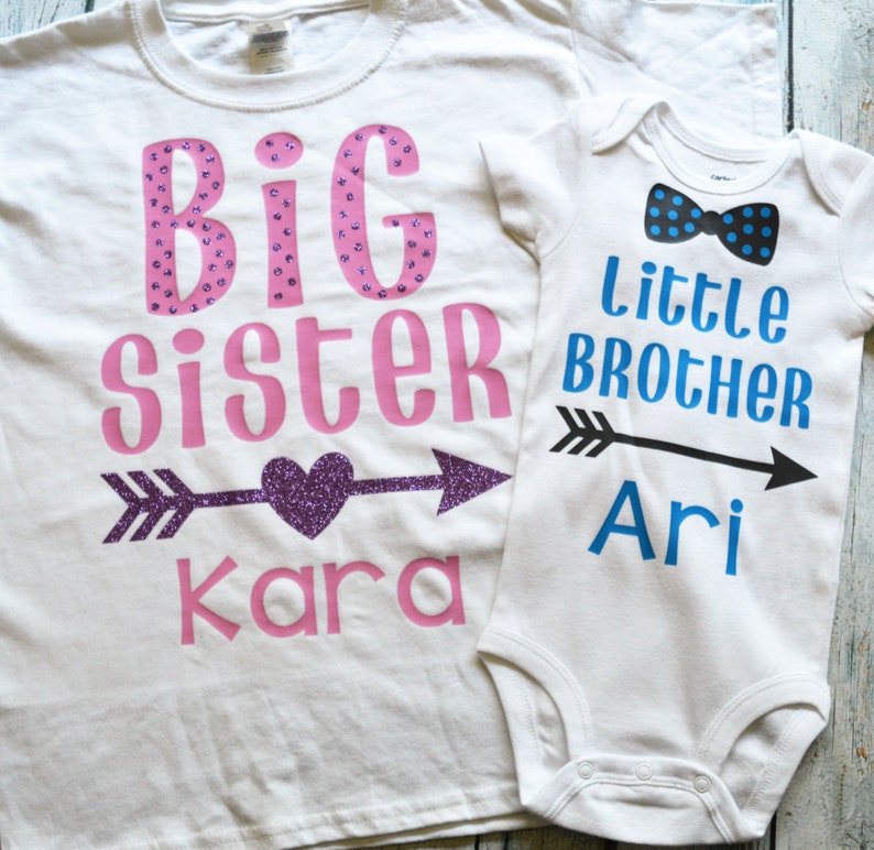 Pu&ograve; includere: Due magliette bianche con un design rosa e blu. La maglietta pi&ugrave; grande dice "Sorella Maggiore Kara" con un cuore glitterato viola e una freccia. La maglietta pi&ugrave; piccola dice "Fratello Minore Ari" con un papillon blu e una freccia.