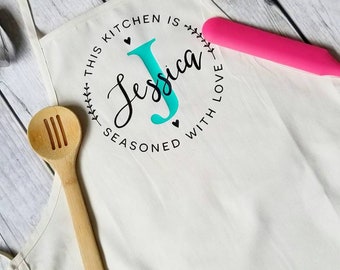 May include: White kitchen apron with a black circle design that says "This kitchen is seasoned with love" and a teal "J" for Jessica.