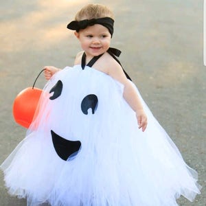 May include: A young child dressed as a ghost for Halloween. The costume is a white tulle dress with a black smiling face on the front. The child is holding an orange pumpkin bucket.