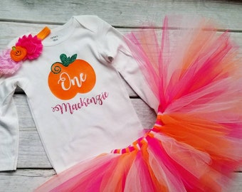 May include: A white long-sleeved onesie with an orange pumpkin design and the text "One Mackenzie". A pink and orange tulle tutu with a white headband with a pink flower and a small orange pumpkin.