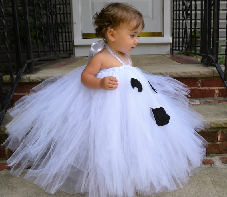 May include: A white tulle tutu with a ghost face design. The ghost has black eyes and a mouth. The tutu is perfect for a Halloween costume.