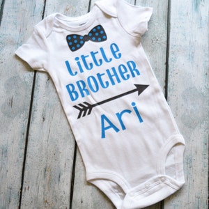 Pu&ograve; includere: Body bianco per neonato con testo blu che recita "Little Brother Ari" con una freccia nera che punta a destra. Un papillon blu e nero &egrave; sopra il testo.